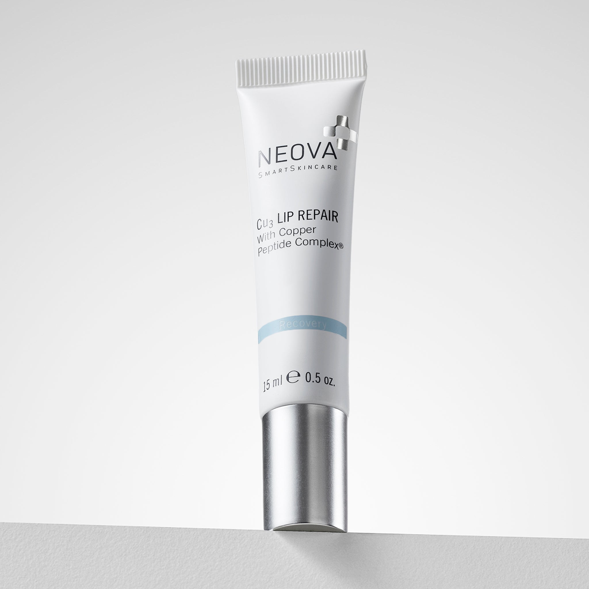 NEOVA Cu3 Lip Repair containing Copper Peptide Complex®, in a small squeeze tube. The front label features a light blue "Recovery" banner. The tube is shown upside down, resting on its flat, brushed silver screw-cap. It contains 0.5 oz. (15 ml) and is set against a clean, bright white background.
