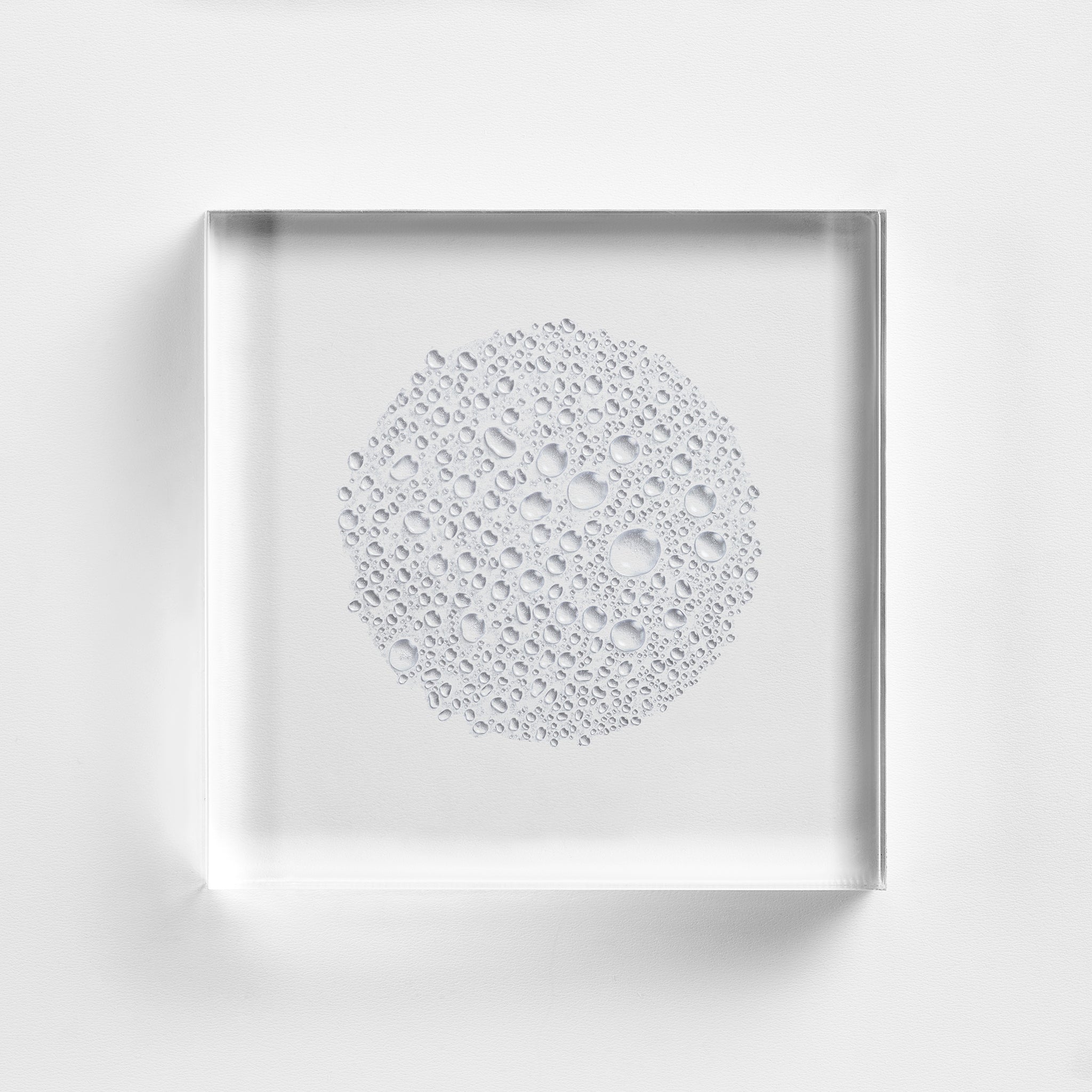 NEOVA Intensive Retinol Spray droplets arranged in a circle on a clear acrylic square. The top-down, macro view shows the transparent droplets are completely clear with a water-like consistency. The composition is brightly lit against a white background, emphasizing the formula's lightweight, aerosol nature.