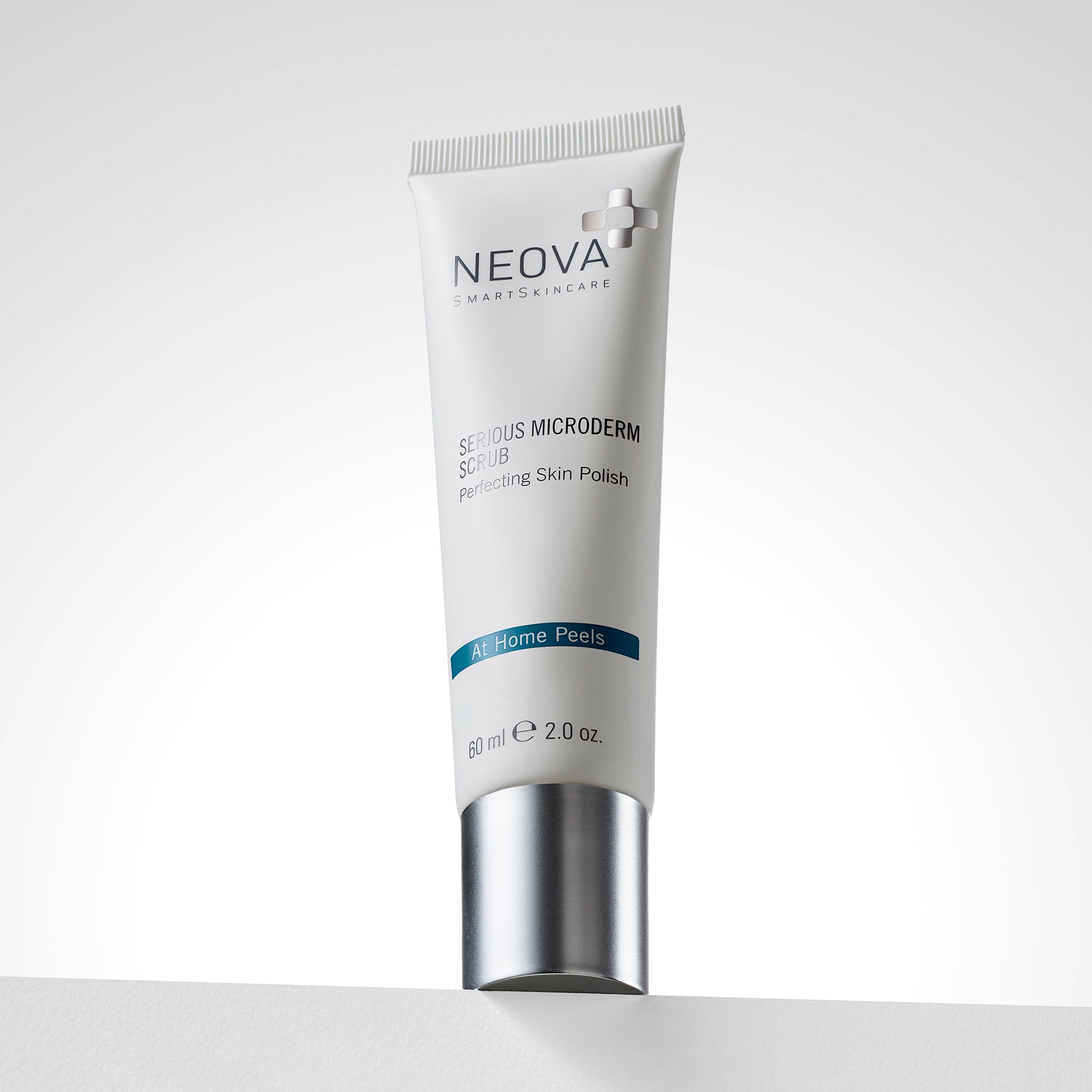 NEOVA Serious Microderm Scrub in a white squeeze tube. The front label describes it as an exfoliant for perfecting skin polish and features a dark blue "At Home Peels" banner. The tube stands upright on its flat, brushed silver screw-cap and contains 2.0 oz (60 ml). It is set on a white ledge against a clean, bright minimalist background.