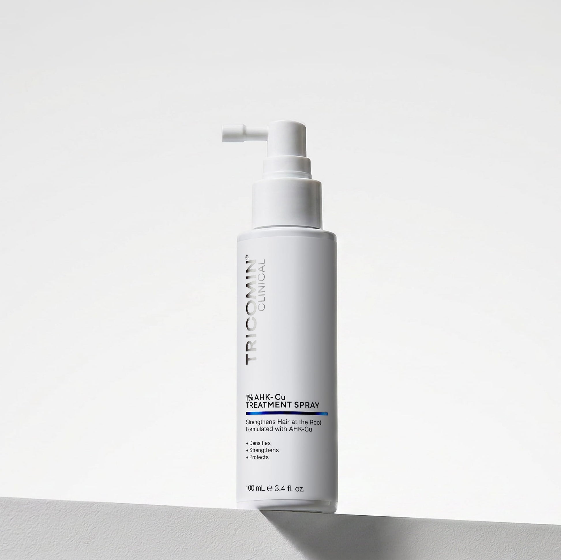 NEOVA Tricomin 1% AHK-Cu Treatment Spray in a white bottle. The label describes notes the hair treatment spray "Strengthens Hair at the Root." and is "Formulated with AHK-Cu" to "Densify", "Strengthen" and "Protect". The bottle features a white spray top and contains 3.4 fl. oz. (100 ml). It is shown standing on a white surface against a clean, bright minimalist background.