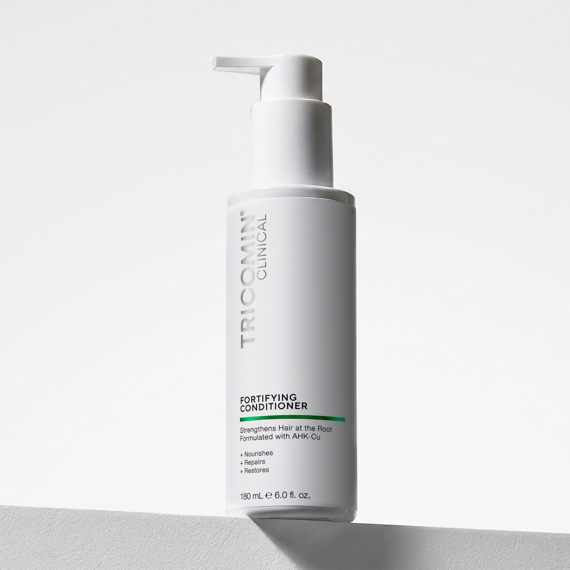 NEOVA Tricomin Fortifying Conditioner in a white bottle. The label describes notes the hair conditioner "Strengthens Hair at the Root." and is "Formulated with AHK-Cu" to "Nourish", "Repair" and "Restore". The bottle features a brushed white pump top and contains 6.0 fl. oz. (180 ml). It is shown standing on a white surface against a clean, bright minimalist background.