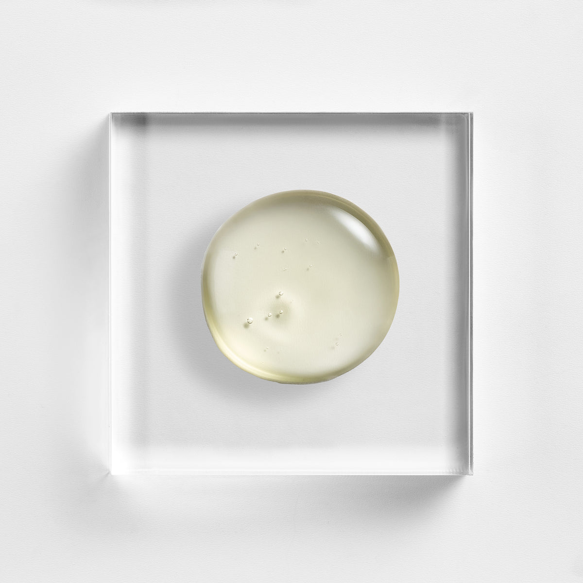 NEOVA Advanced HA serum centered on a clear acrylic square. The top-down macro view shows the serum is a pale, golden-tinted gel containing tiny, suspended oxygen bubbles, suggesting a lightweight and hydrating formula. The shot is well-lit against a textured white backdrop, highlighting the serum's clarity and viscosity.