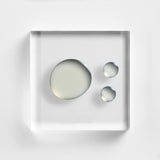 NEOVA C 20+ serum on a clear acrylic square. There is one large, circular drop and two smaller droplets to the right in the top-down view. The liquid is transparent with a very faint, pale yellow tint and a smooth, water-like viscosity.