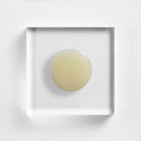 NEOVA Cell Renewal serum on a clear acrylic square. There is one large, circular drop in the center of the top-down view. The liquid is transparent with a very faint, pale yellow tint and a smooth, water-like viscosity.