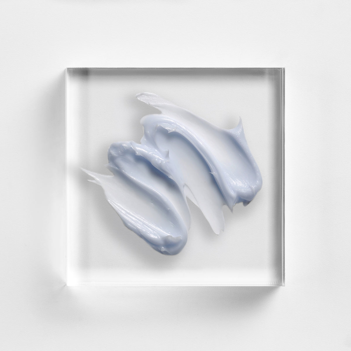 NEOVA Clear Relief texture a clear acrylic square. The top-down macro view shows the product is lavender-grey in color with a creamy texture. It is centered against a textured white background with soft, clean lighting.
