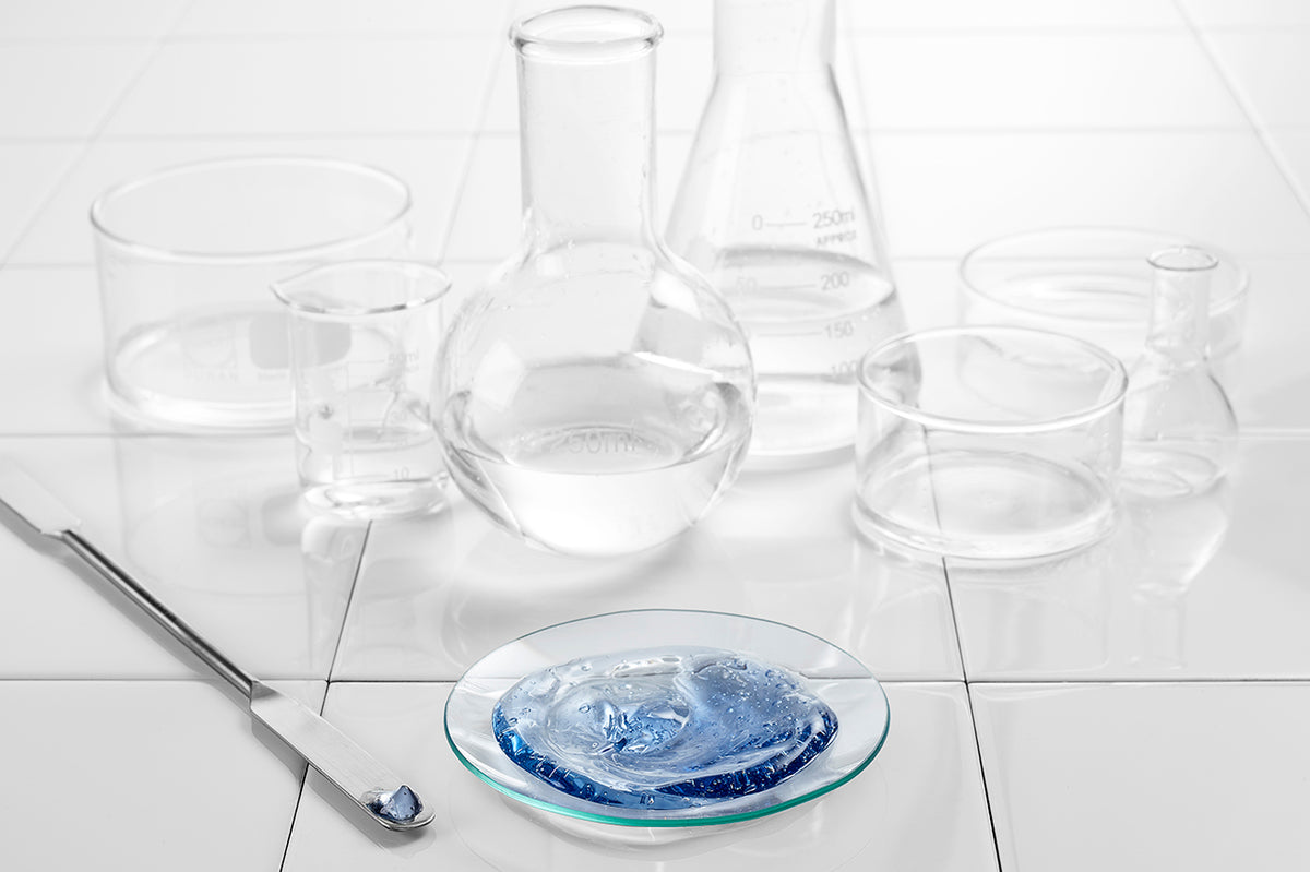 NEOVA Copper Peptide Science - Clear glass laboratory equipment on a white tiled surface with a blue copper peptide facial mask in a glass dish in the foreground.