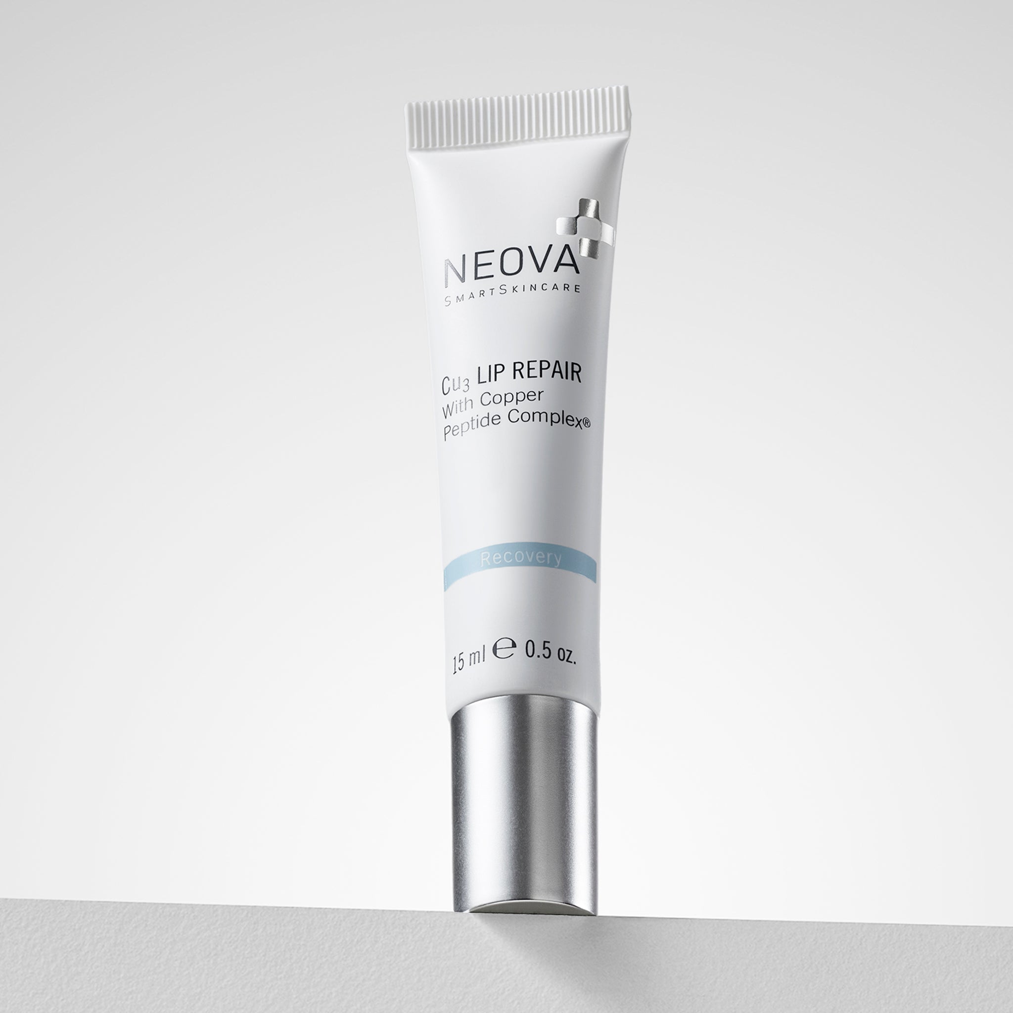 NEOVA Cu3 Lip Repair containing Copper Peptide Complex®, in a small squeeze tube. The front label features a light blue "Recovery" banner. The tube is shown upside down, resting on its flat, brushed silver screw-cap. It contains 0.5 oz. (15 ml) and is set against a clean, bright white background.