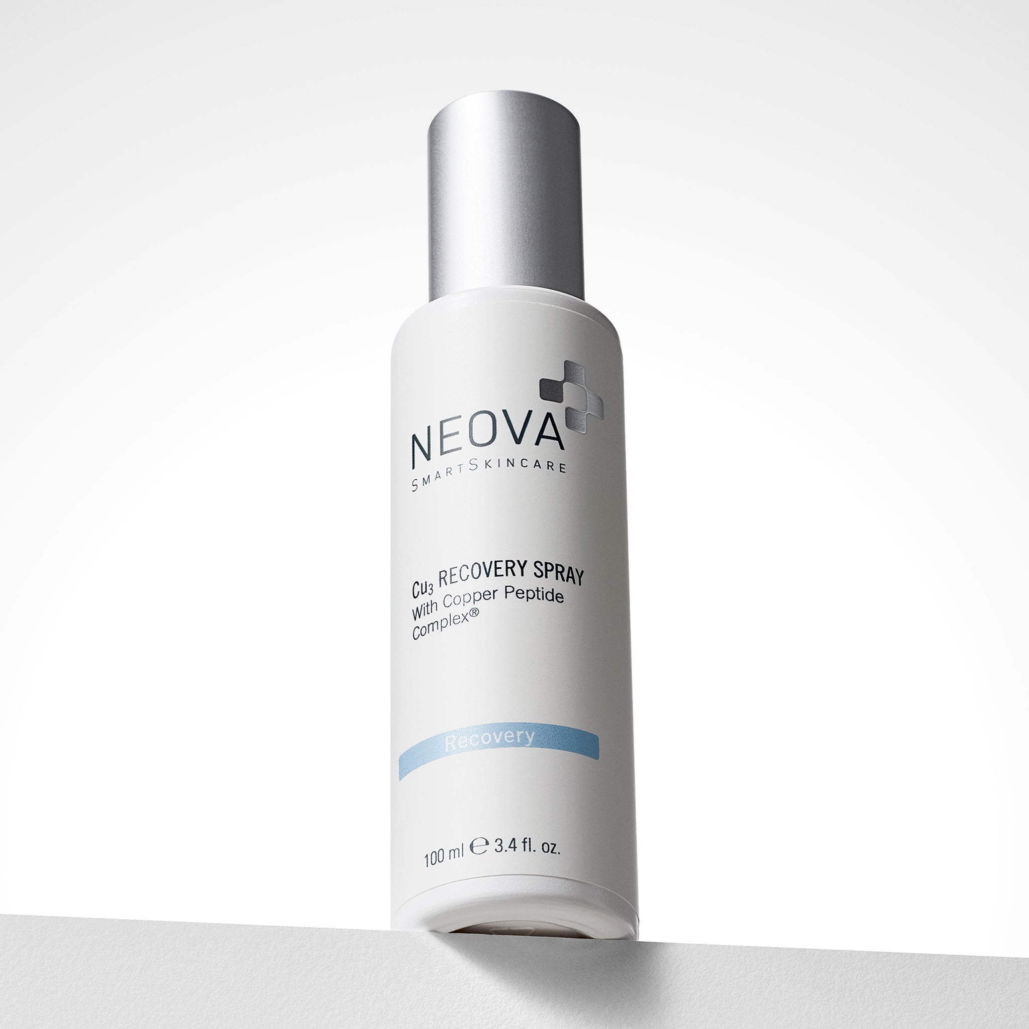 NEOVA Cu3 Recovery Spray, formulated with Copper Peptide Complex®, in a white cylindrical spray bottle. The front label features a light blue "Recovery" banner. The bottle is capped with a brushed silver lid and contains 3.4 fl. oz. (100 ml). It is photographed from a low-angle perspective, standing on a white ledge against a clean, bright background with soft gradients.