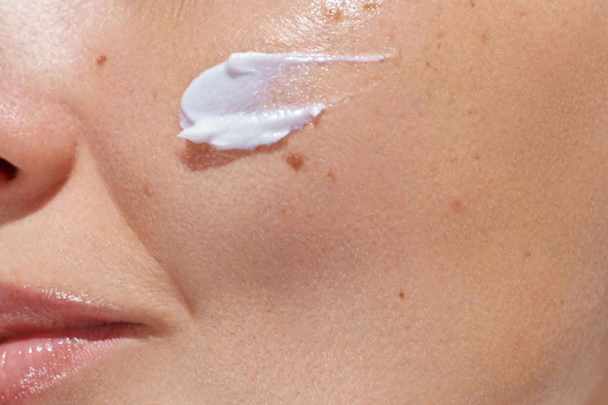 "NEOVA Daily Moisture is a clinical hydrating face moisturizer, shown in this close-up application shot where a rich, pale grey-purple swatch is being blended onto a woman's cheek by her fingertips, delivering Copper Peptides and DNA Repair Enzymes for skin revitalization.