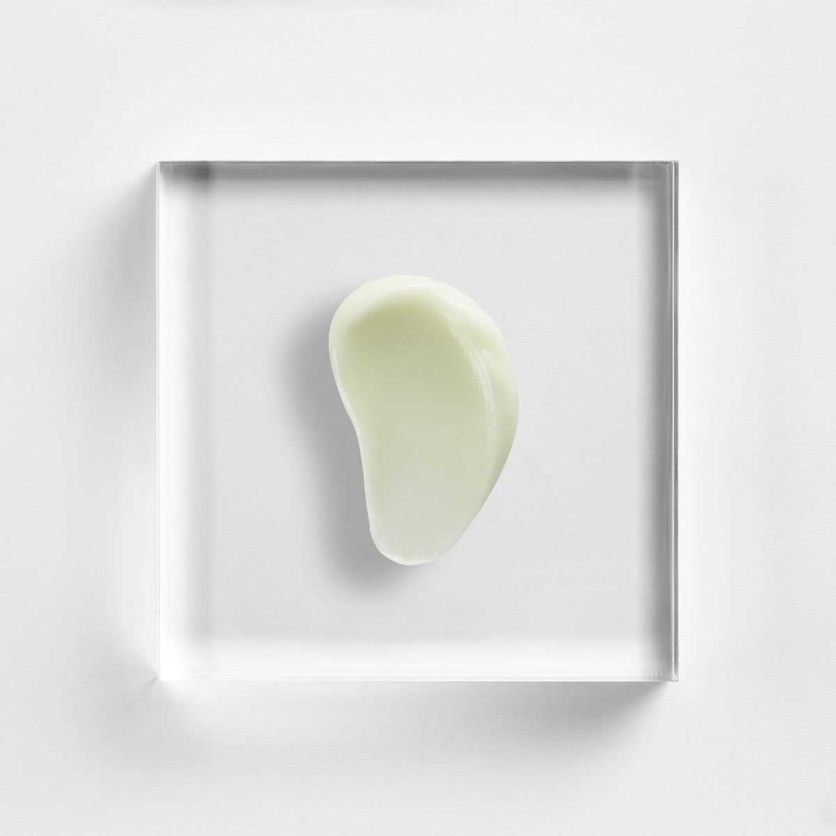 NEOVA Dual Matrix serum on a clear acrylic square. The top-down macro view of the serum shows it has a smooth, semi-opaque, pale yellow or cream-colored appearance with a slightly glossy finish. The composition is centered and brightly lit against a white background, showcasing the formula's lightweight, gel-like consistency.