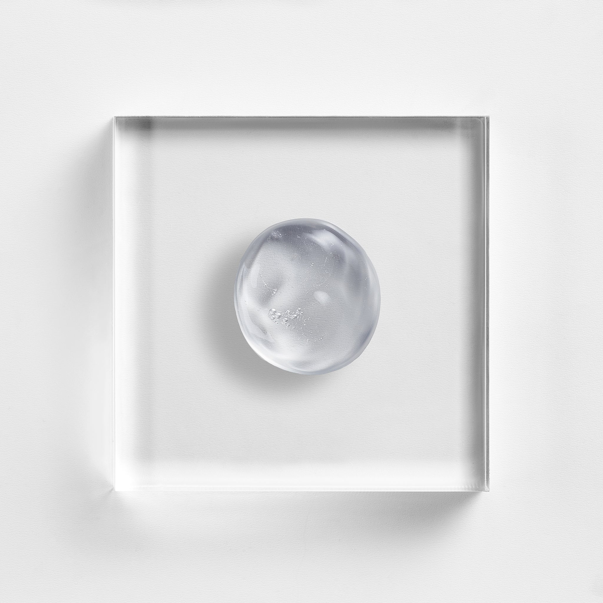 NEOVA Herbal Wash on a clear acrylic block. The top-down, macro composition shows the large, circular drop of cleanser is a transparent gel with a smooth, viscous consistency and a few tiny, suspended bubbles. The shot is brightly lit against a textured white background, emphasizing the gel's clarity.