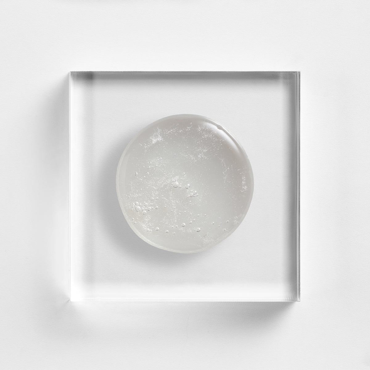 NEOVA Microfoliant Cleanser on a clear acrylic square. The top-down, macro view shows the cleanser is an opaque white gel with a visible, fine-grained texture from the micro-exfoliants. The composition is centered against a minimalist white background to highlight its creamy yet gritty consistency.