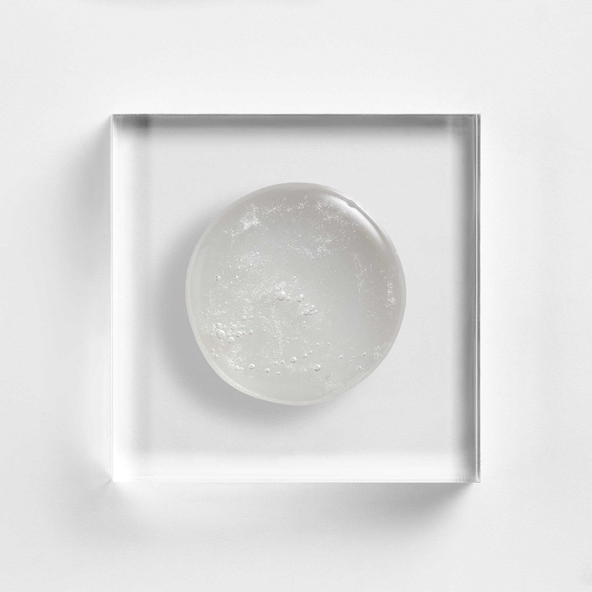 NEOVA Microfoliant Cleanser on a clear acrylic square. The top-down, macro view shows the cleanser is an opaque white gel with a visible, fine-grained texture from the micro-exfoliants. The composition is centered against a minimalist white background to highlight its creamy yet gritty consistency.