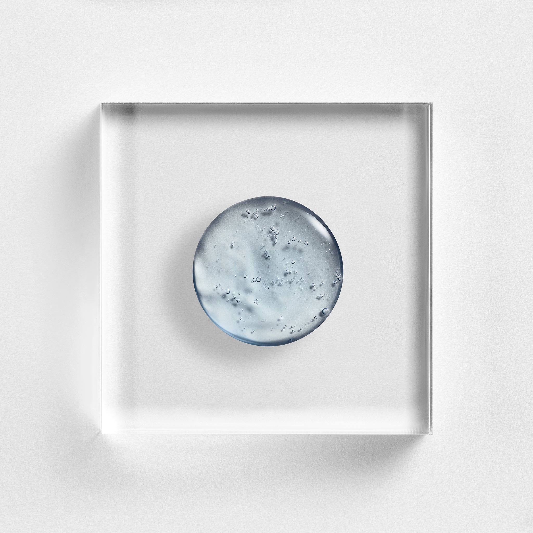 NEOVA Power Defense serum on a clear acrylic square. The top-down, square macro view shows the serum has a distinct, pale sky-blue tint and a smooth, lightweight gel-cream consistency with a glossy finish. It is centered and brightly lit against a textured white background, highlighting the formula's unique color and fluid texture.