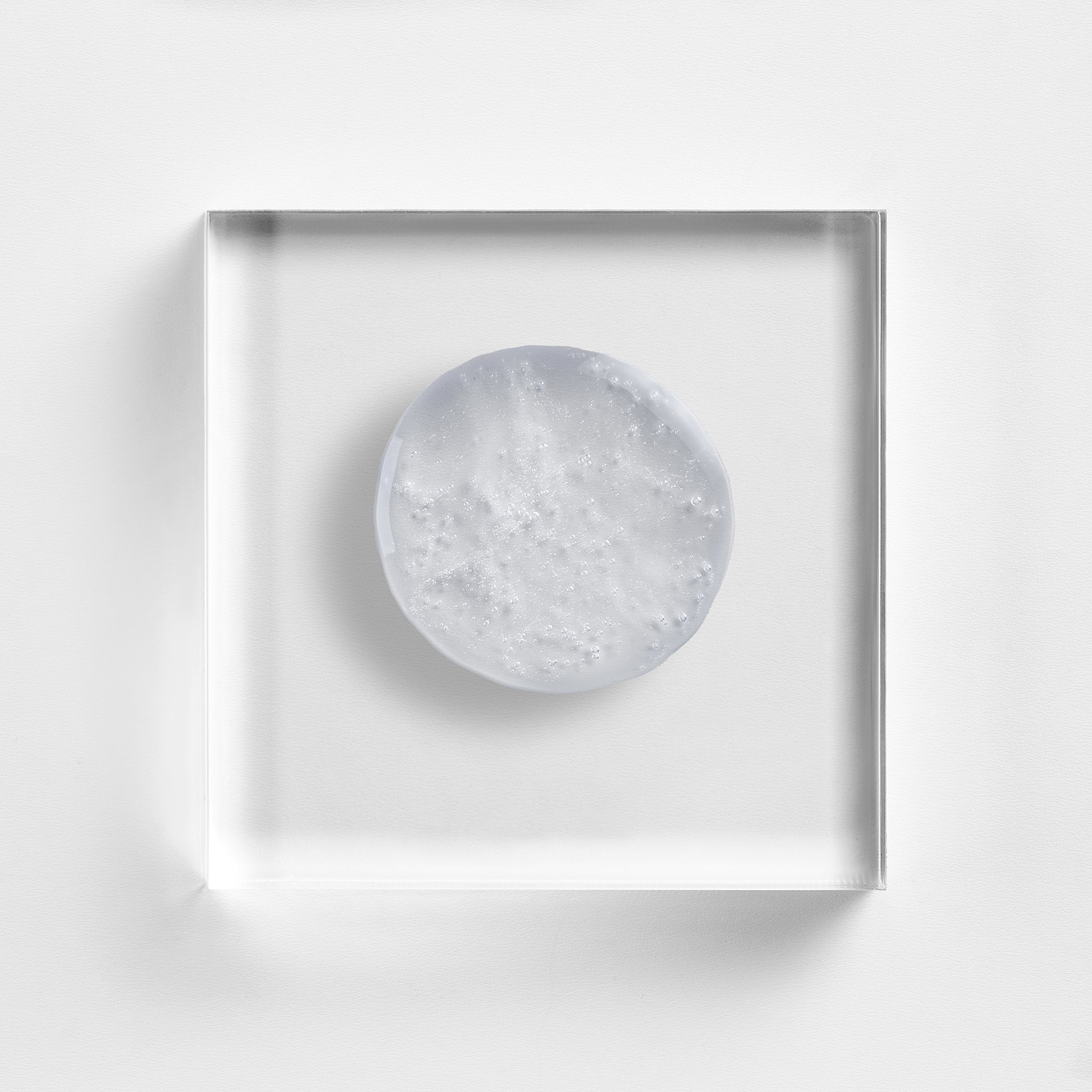 NEOVA Radiant Wash on a clear acrylic square. The top-down, macro composition shows the large, circular drop of cleanser is transparent, colorless gel with a smooth, viscous consistency and a few small, suspended oxygen bubbles. It is centered and brightly lit against a textured white background, highlighting its clean and fluid appearance.