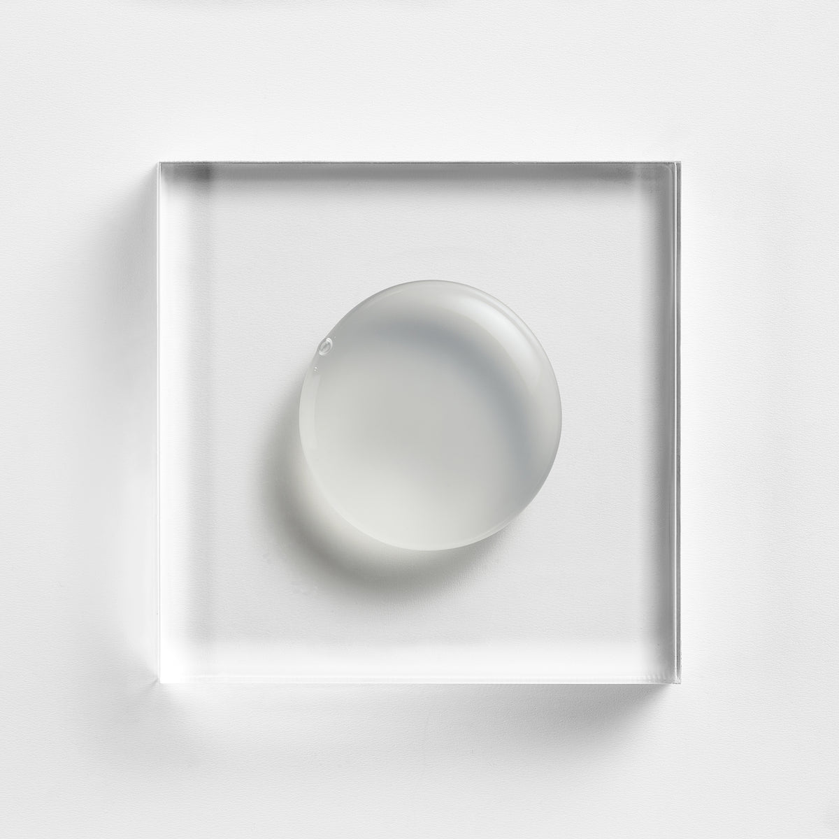 NEOVA Serious Reveal Exfoliator on a clear acrylic square. The top-down, square macro view shows the product is a smooth, opaque white cream with a lightweight, fluid consistency and a soft glossy finish. It is centered and brightly lit against a textured white background, highlighting its creamy and refined texture.