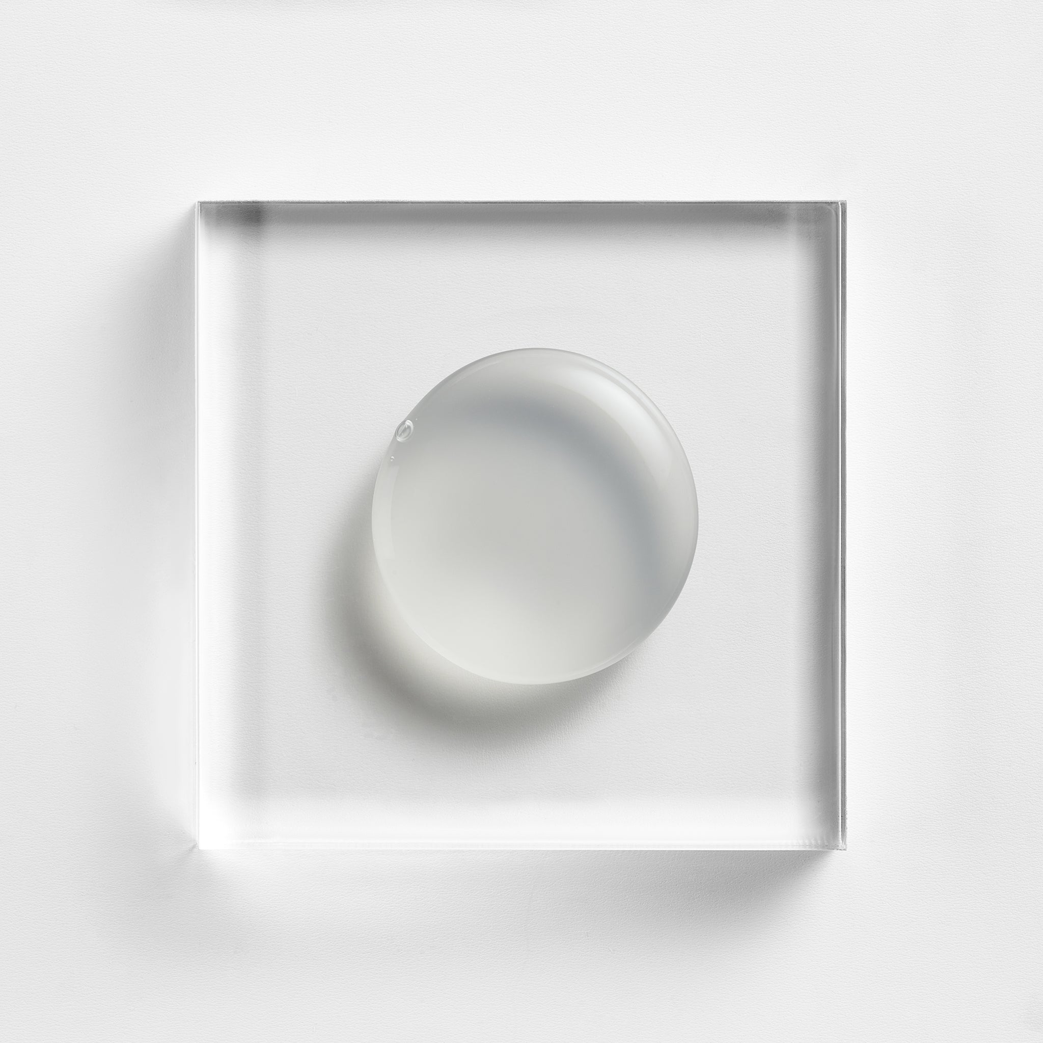 NEOVA Serious Reveal Exfoliator on a clear acrylic square. The top-down, square macro view shows the product is a smooth, opaque white cream with a lightweight, fluid consistency and a soft glossy finish. It is centered and brightly lit against a textured white background, highlighting its creamy and refined texture.
