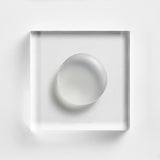 NEOVA Serious Reveal Exfoliator on a clear acrylic square. The top-down, square macro view shows the product is a smooth, opaque white cream with a lightweight, fluid consistency and a soft glossy finish. It is centered and brightly lit against a textured white background, highlighting its creamy and refined texture.