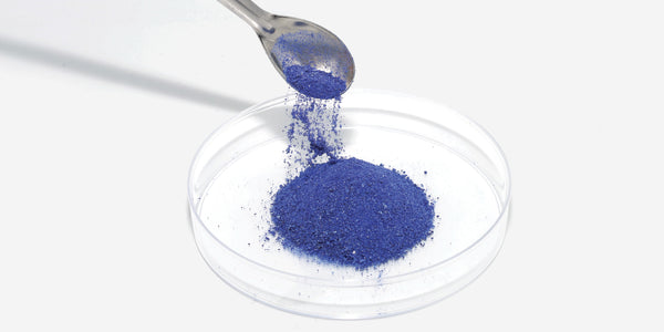 NEOVA - Timeline - 1980 - Blue powder containing Copper Peptides being poured from a spoon into a clear glass dish on a white background