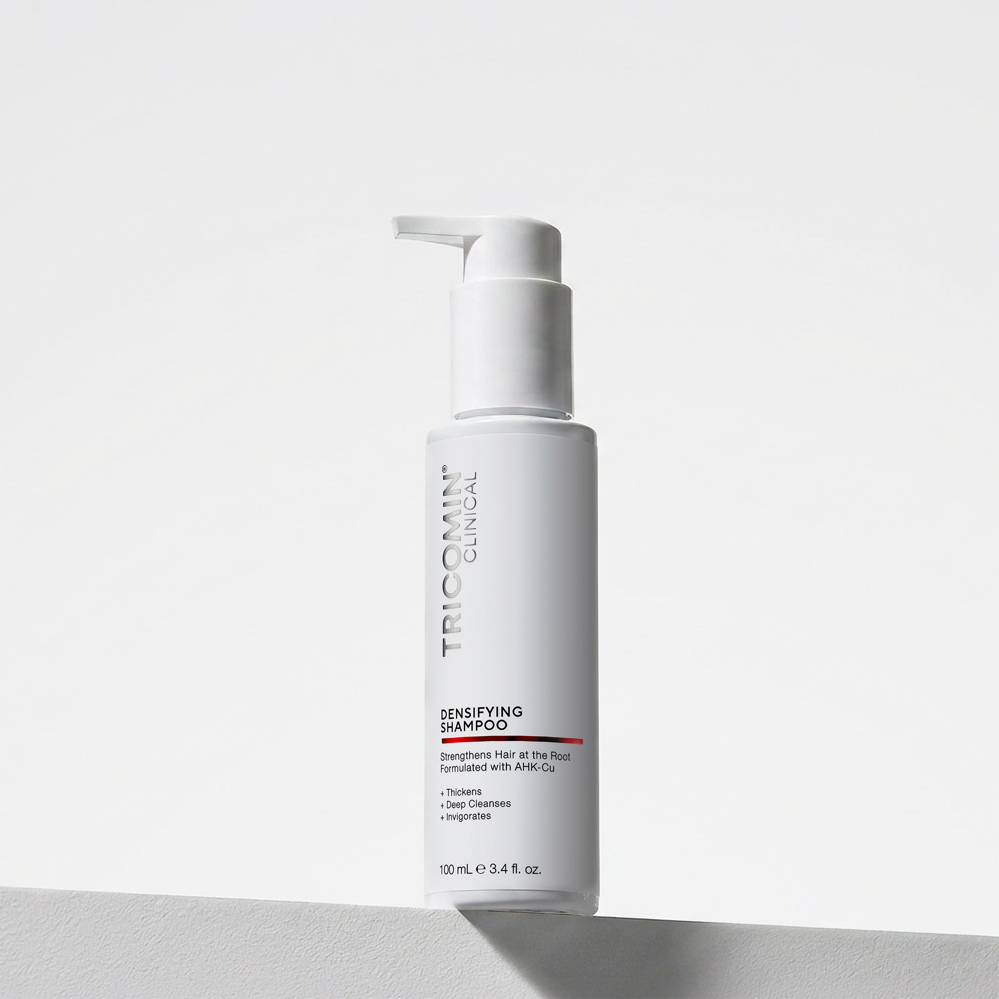 NEOVA Tricomin Densifying Shampoo 100ml in a white bottle. The label describes notes the shampoo "Strengthens Hair at the Root." and is "Formulated with AHK-Cu" to "Thicken", "Deep Cleanse" and "Invigorate". The bottle features a white pump top and contains 3.4 fl. oz. (100 ml). It is shown standing on a white surface against a clean, bright minimalist background.