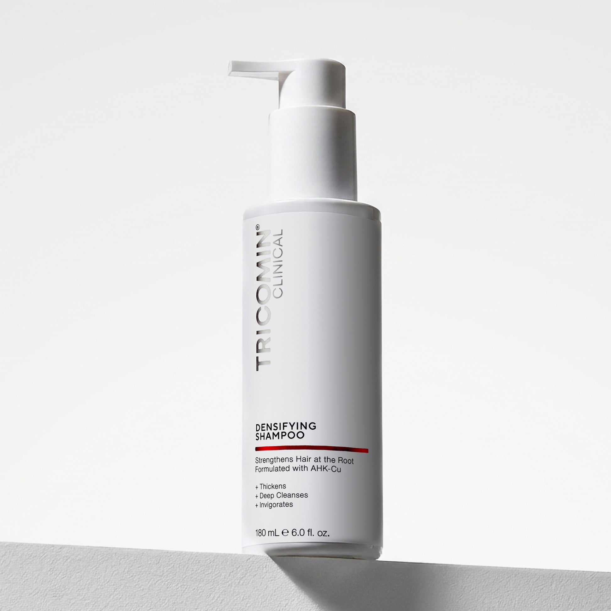 NEOVA Tricomin Densifying Shampoo in a white bottle. The label describes notes the shampoo "Strengthens Hair at the Root." and is "Formulated with AHK-Cu" to "Thicken", "Deep Cleanse" and "Invigorate". The bottle features a white pump top and contains 6.0 fl. oz. (180 ml). It is shown standing on a white surface against a clean, bright minimalist background.